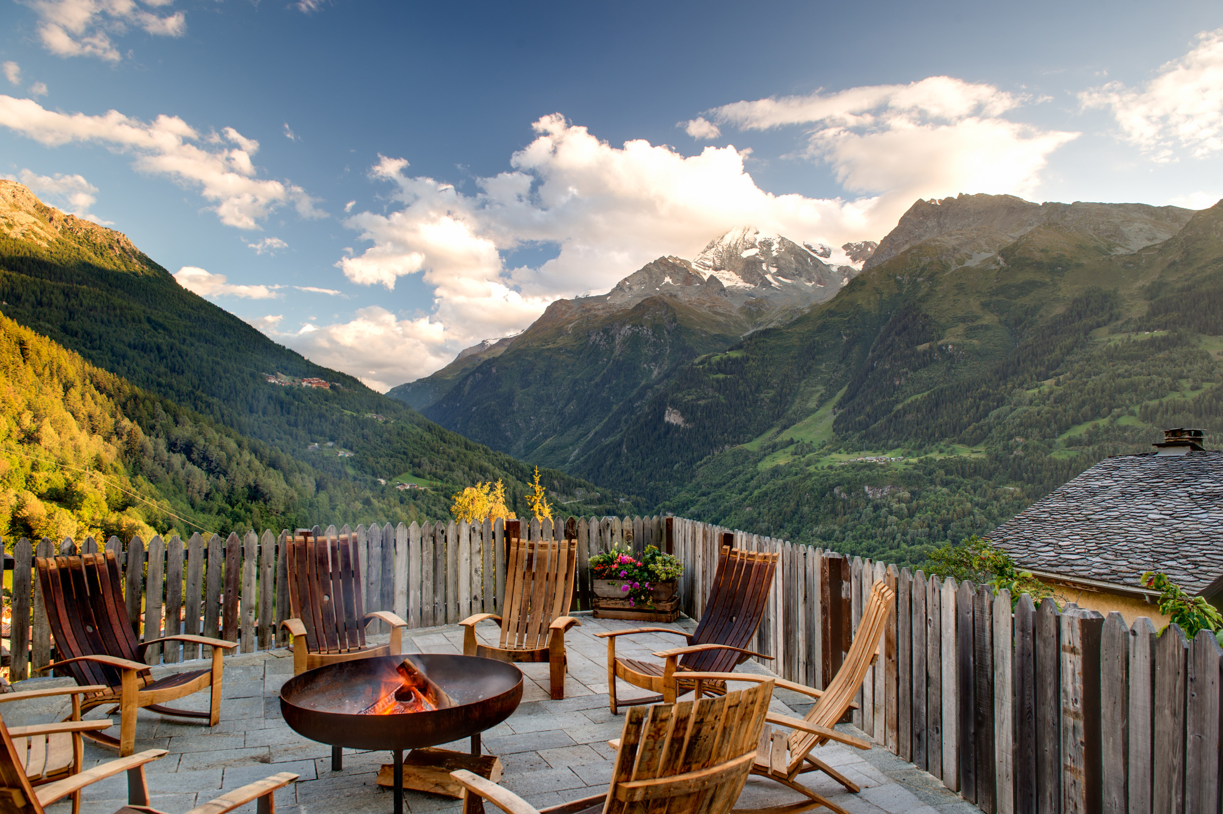 Exterior porch view overlooking the Alps on a bright summer day, with a fire and deck chairs in the foreground.