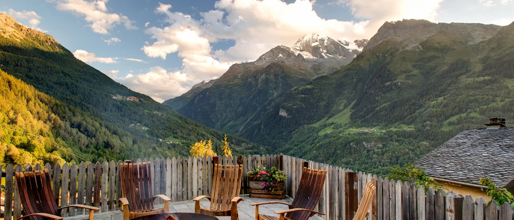 Exterior porch view overlooking the Alps on a bright summer day, with a fire and deck chairs in the foreground.