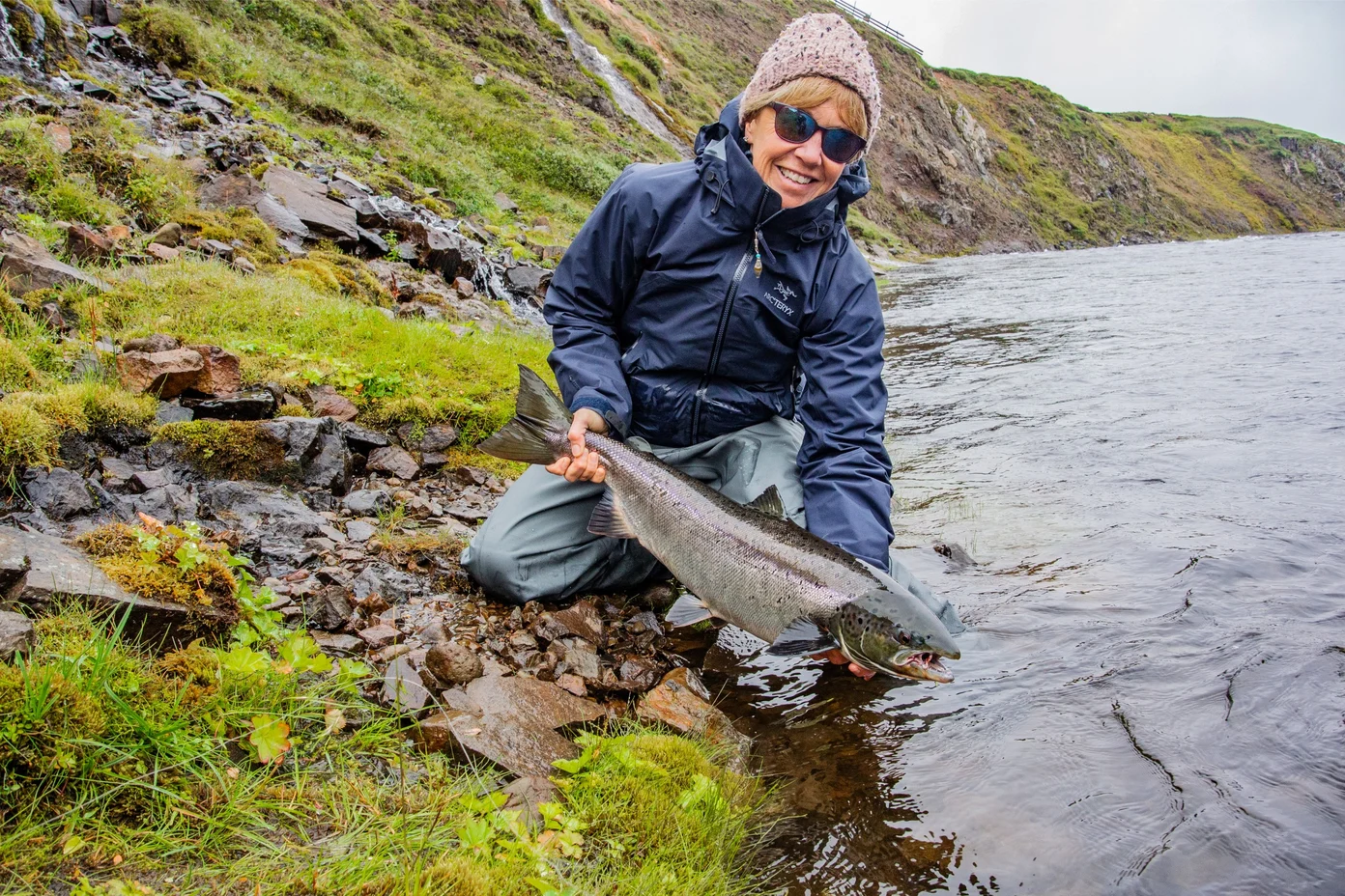 A woman holding an Atlantic char at the edge of a river.