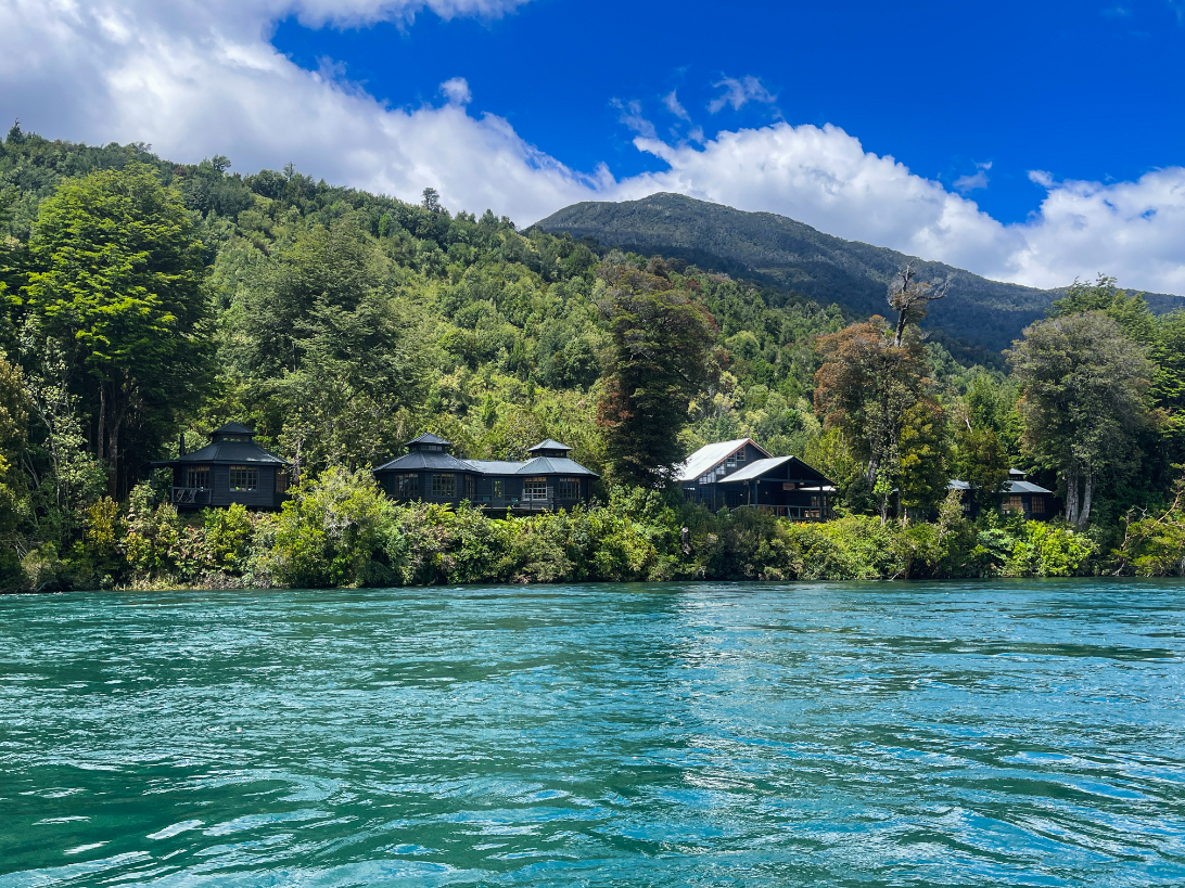 A lodge sits on a river bank with lush trees and greenery surrounding the lodge and bright blue river waters in the foreground on a bright summer day.