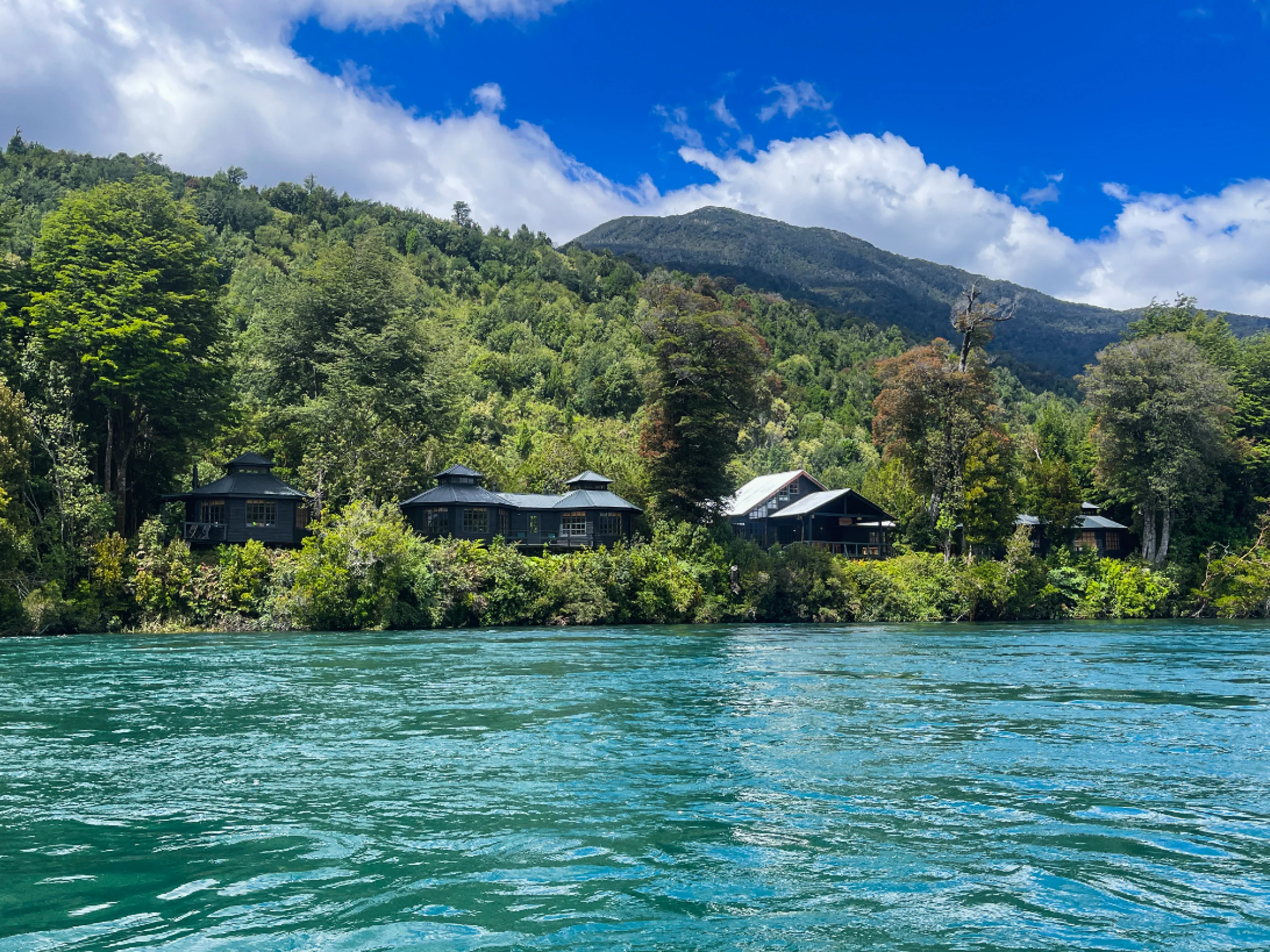 A lodge sits on a river bank with lush trees and greenery surrounding the lodge and bright blue river waters in the foreground on a bright summer day.