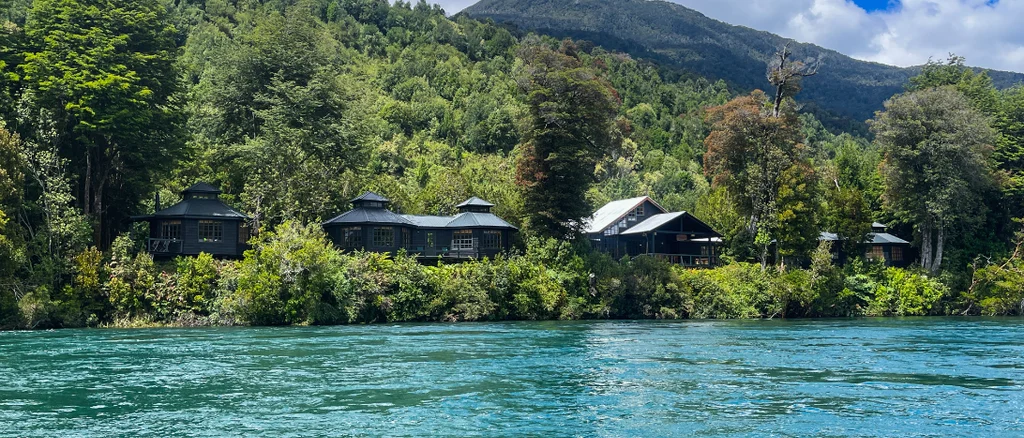 A lodge sits on a river bank with lush trees and greenery surrounding the lodge and bright blue river waters in the foreground on a bright summer day.