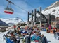A large group of skiers hanging out at the bottom of a ski lift at a lodge.