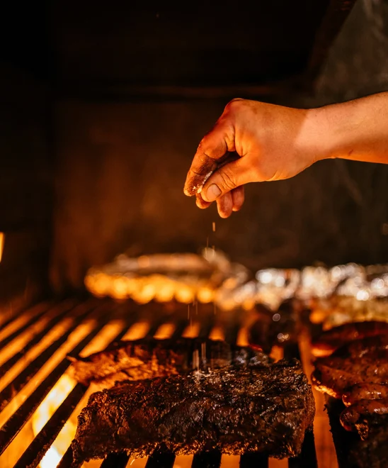 A chef's hand sprinkling salt on top of a large steak on the grill.