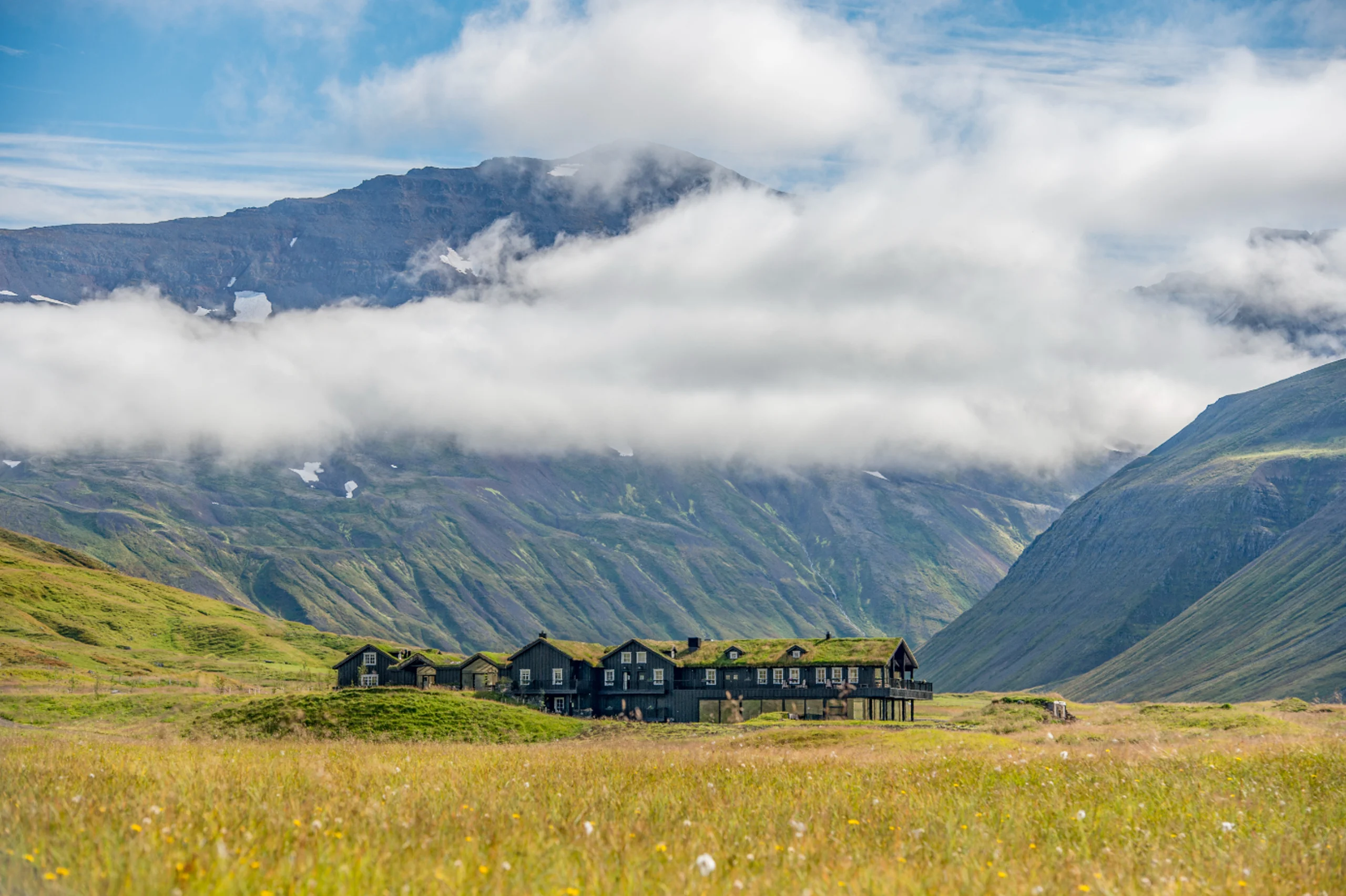 A large lodge is seen with clouds above, mountains behind on a summer day. The lodge has a dark exterior with a green roof. There is a large field in the foreground.