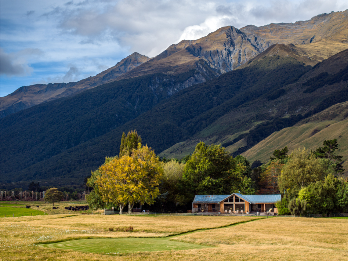 A lodge sits in the center of a field with mountains behind it. Trees surround the lodge on a sunny day.