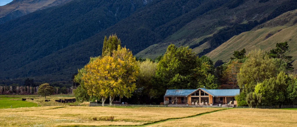 A lodge sits in the center of a field with mountains behind it. Trees surround the lodge on a sunny day.