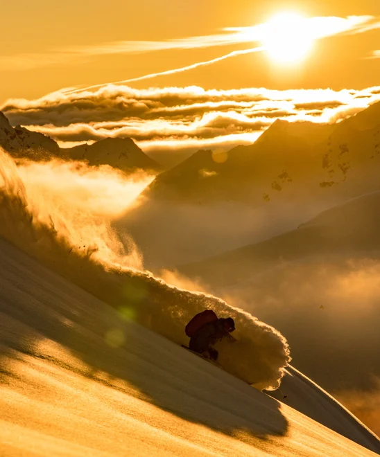 A person skiing in a cloud of snow above the clouds in French Alps at sunset.