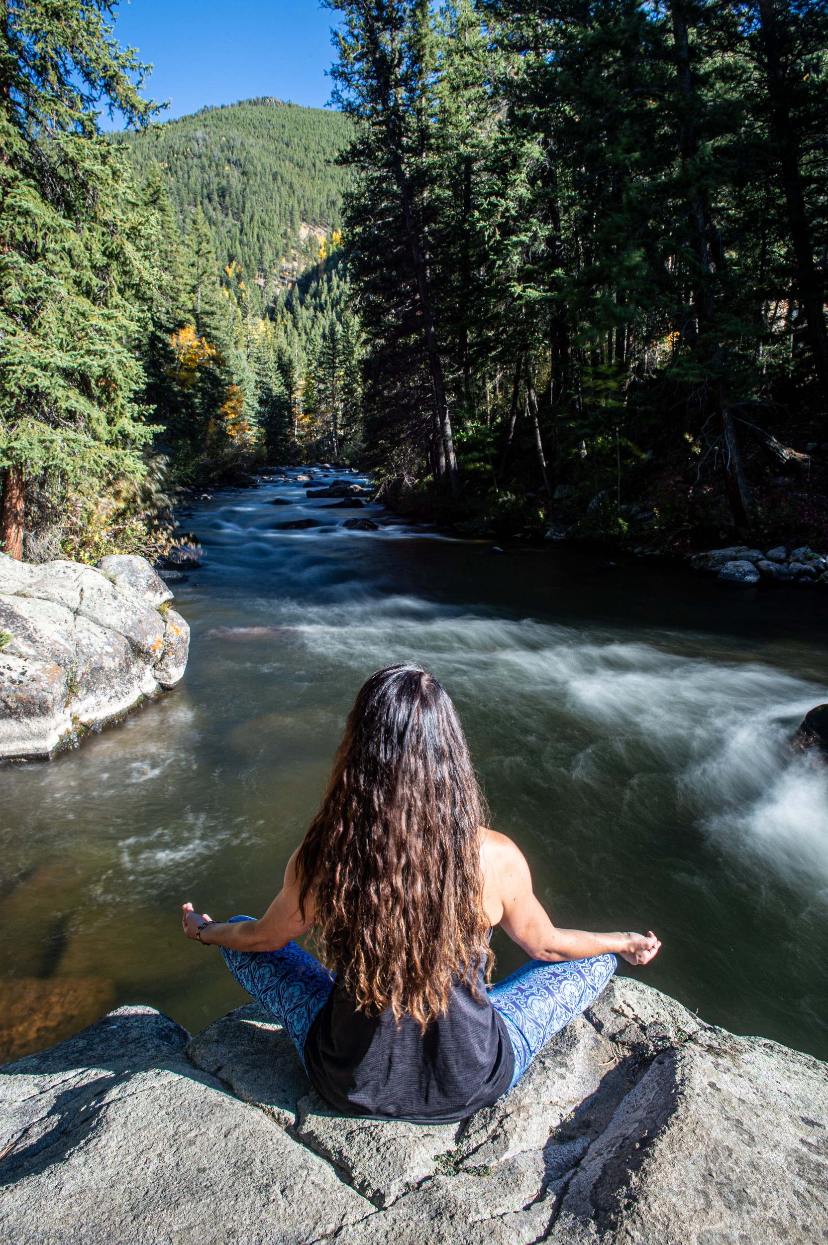 A person sitting meditating on a boulder at river's edge.