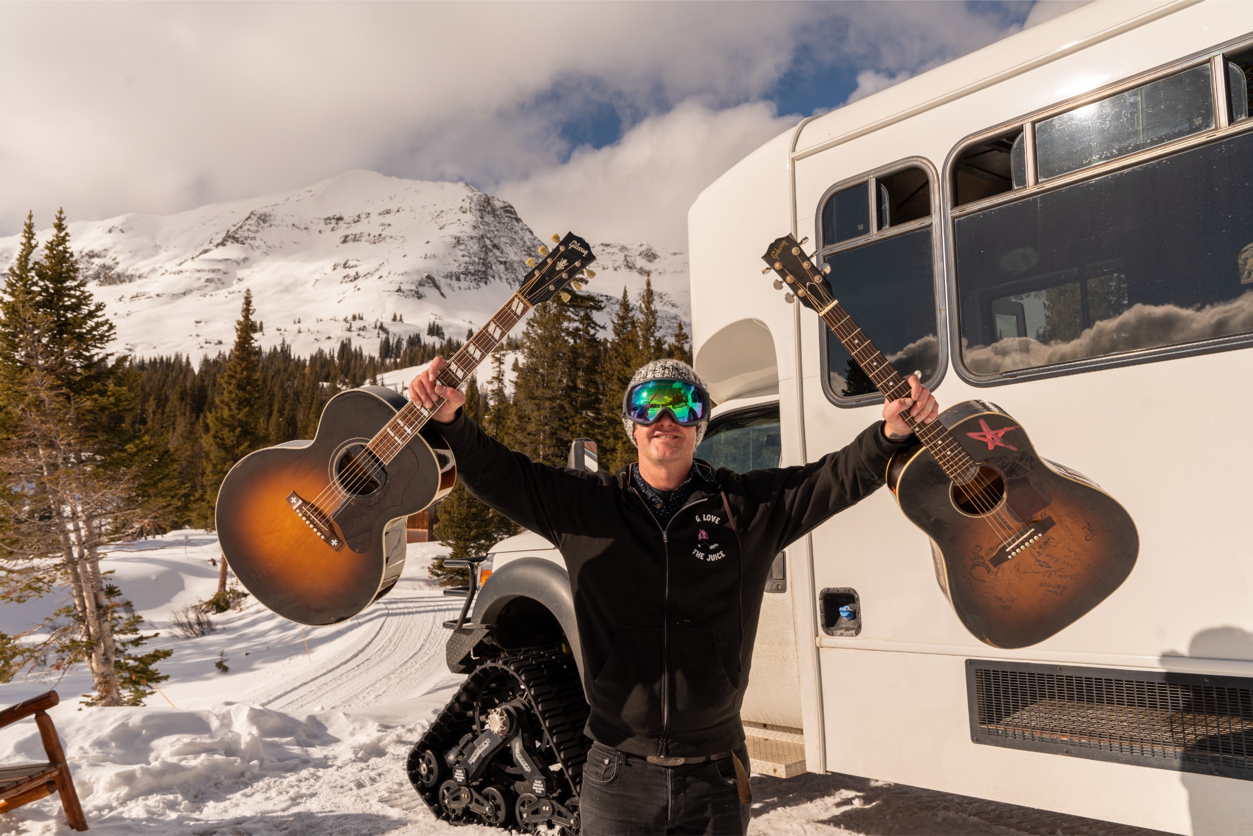 A musician holding two acoustic guitars outside next to a cat ski.