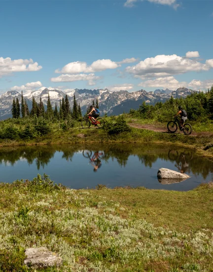 Two people mountain biking around a pond on a small mountain trail.