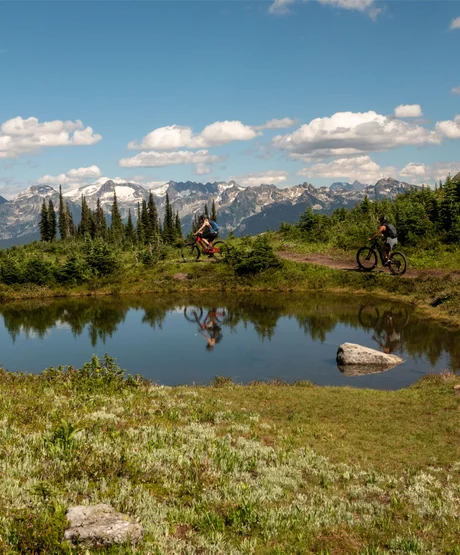 Two people mountain biking around a pond on a small mountain trail.