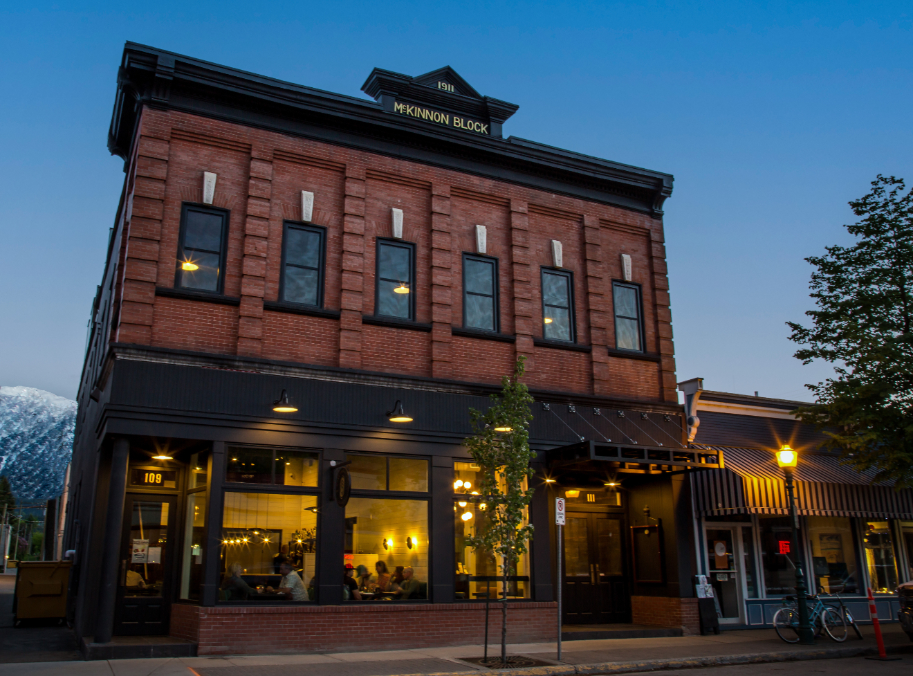 The historical McKinnong building in Revelstoke is seen on a winter night with the lights from the restaurant glowing on the first floor and a snow covered mountain in the background.