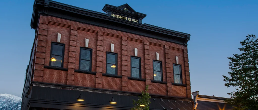 The historical McKinnong building in Revelstoke is seen on a winter night with the lights from the restaurant glowing on the first floor and a snow covered mountain in the background.