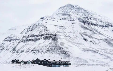 A large dark lodge sits at the base of a snow covered mountain. The lodge itself is snow covered with snow all around.