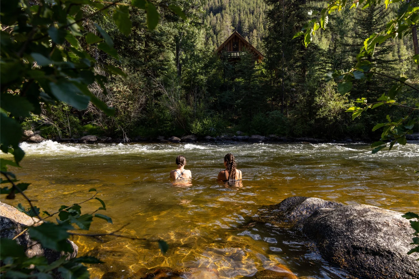 Two people swimming in Taylor River near the lodge, surrounded by trees.