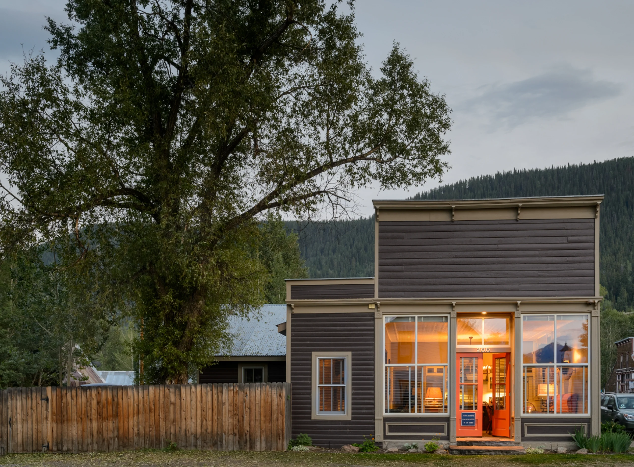A historical western building sits at the end of the lane, with the light on and red door open. It is a warm and inviting lodge exterior on a summer evening with trees and mountains in the background.