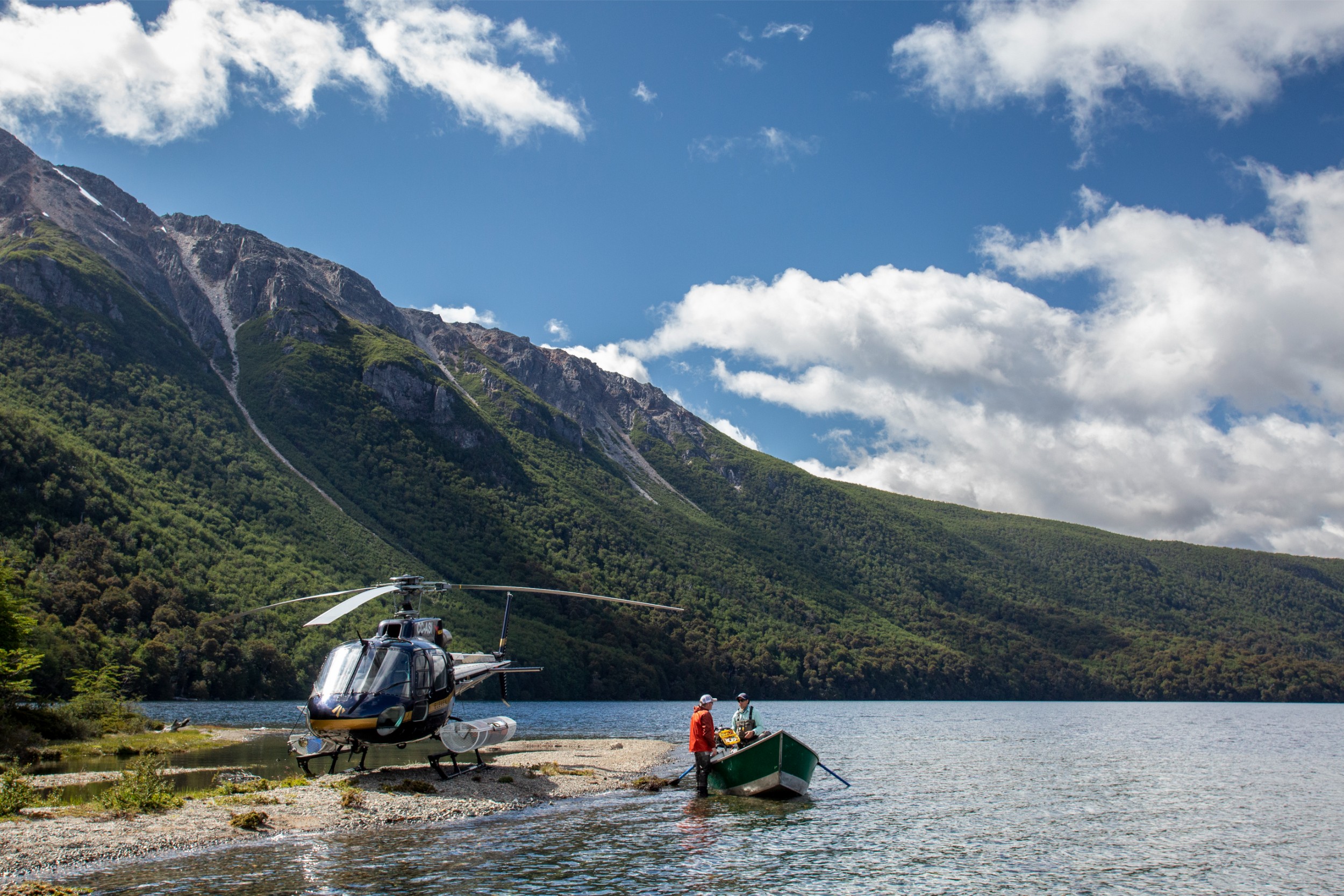 A helicopter parked in a remote river valley with two fishers on a small boat.