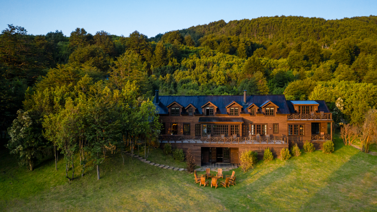 A warm colored wood lodge sits at the base of a mountain, with a large lawn and chairs in front on a bright summer day.