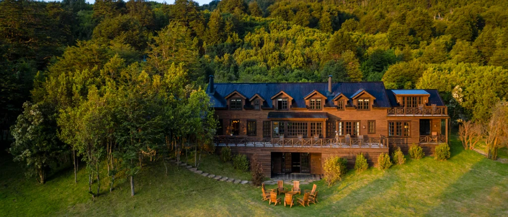 A warm colored wood lodge sits at the base of a mountain, with a large lawn and chairs in front on a bright summer day.