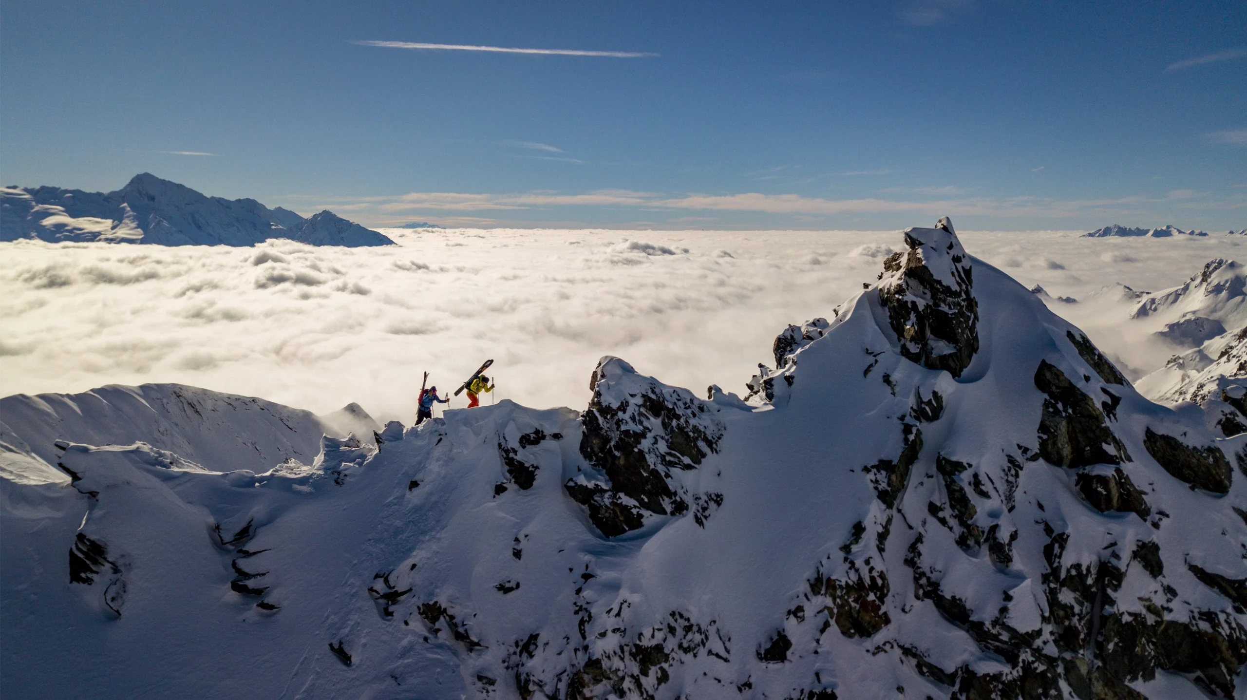 Two skiers walking across the ridge of a mountain above the clouds.