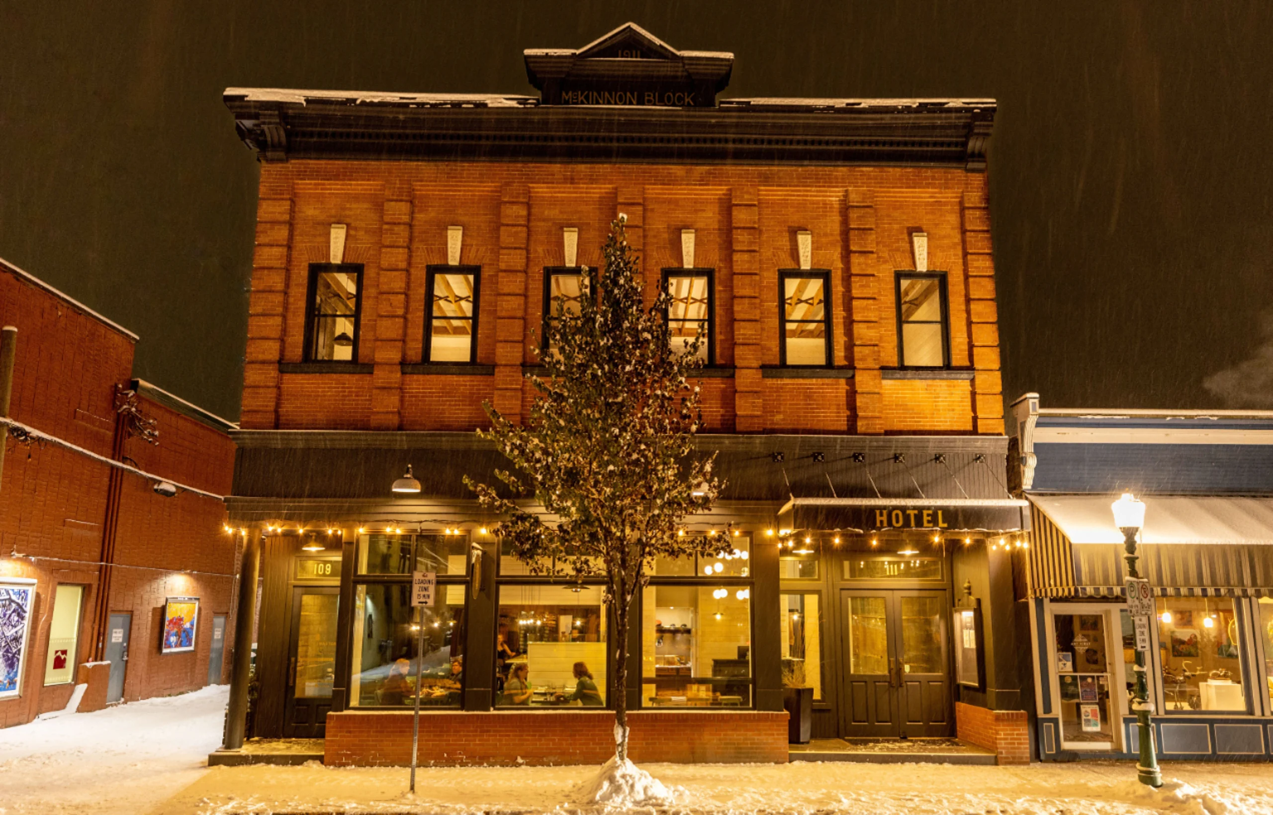 A snow covered brike building is seen in historic downtown Revelstoke on a winter night. A warm glow comes from the lodging and restaurant inside.